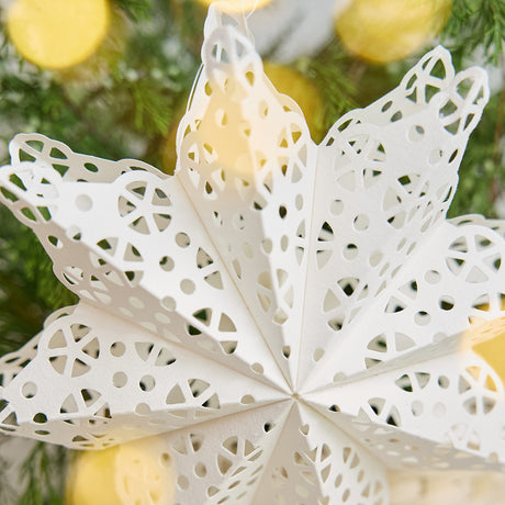 Craftspring’s large white Paper Snowflakes Christmas Ornament with intricate cutout patterns, displayed on snow in front of a basket of evergreens, adding elegant holiday charm, ethically handmade by artisans from recycled cotton waste.