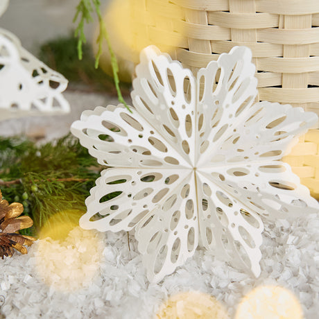Craftspring’s smaller white Paper Snowflakes Christmas Ornament with intricate cutout patterns, displayed on snow in front of a basket of evergreens, adding delicate holiday charm, ethically handmade by artisans from recycled cotton waste.