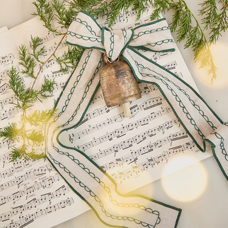 Craftspring’s Embossed Bell with Green Ribbon — handmade using a special process and hand-tuned for a clear ring, with a linen ribbon embroidered in green — displayed on sheet music with evergreens for a festive holiday scene, ethically crafted by artisans.