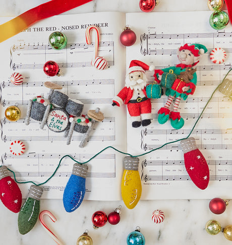 Christmas Drum Set Ornament, Elf with Gifts Ornament, and Little Santa with Gift displayed together on sheet music with shiny baubles and Christmas candy — handmade felt Christmas ornaments from Craftspring, richly detailed with embroidery and festive accents, ethically crafted by artisans using sustainable wool for joyful holiday gifting.