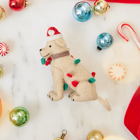 Craftspring’s Yellow Lab in Christmas Lights Ornament — felt dog wrapped in Christmas lights and wearing a Santa hat — styled among holiday baubles and candy canes, evoking festive warmth, playful holiday charm, and heirloom-quality gifting, ethically crafted by artisans using sustainable wool.