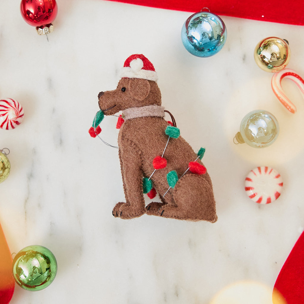 Craftspring’s Chocolate Lab in Christmas Lights Ornament — felt dog wrapped in Christmas lights and wearing a Santa hat — styled among holiday baubles and candy canes, evoking festive warmth, playful holiday charm, and heirloom-quality gifting, ethically crafted by artisans using sustainable wool.