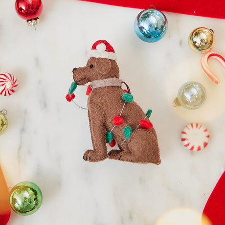 Craftspring’s Chocolate Lab in Christmas Lights Ornament — felt dog wrapped in Christmas lights and wearing a Santa hat — styled among holiday baubles and candy canes, evoking festive warmth, playful holiday charm, and heirloom-quality gifting, ethically crafted by artisans using sustainable wool.