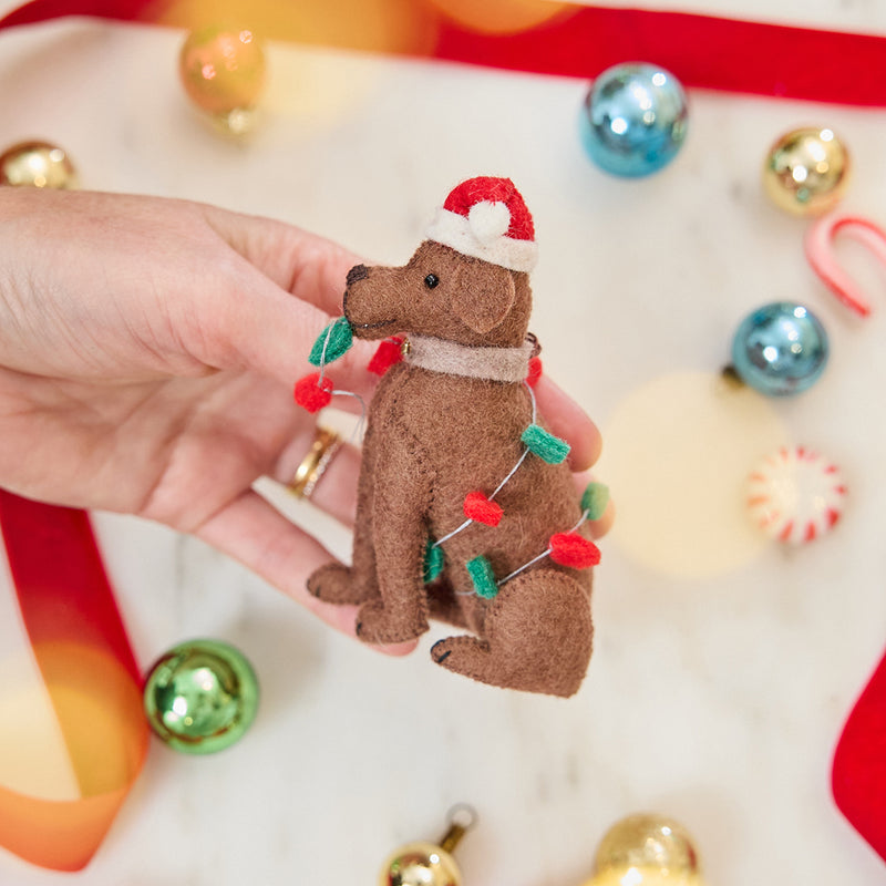 Craftspring’s Chocolate Lab in Christmas Lights Ornament — felt dog wrapped in Christmas lights and wearing a Santa hat — held in hand over holiday baubles, capturing playful charm, festive warmth, and heirloom-quality Christmas gifting, ethically crafted by artisans using sustainable wool.