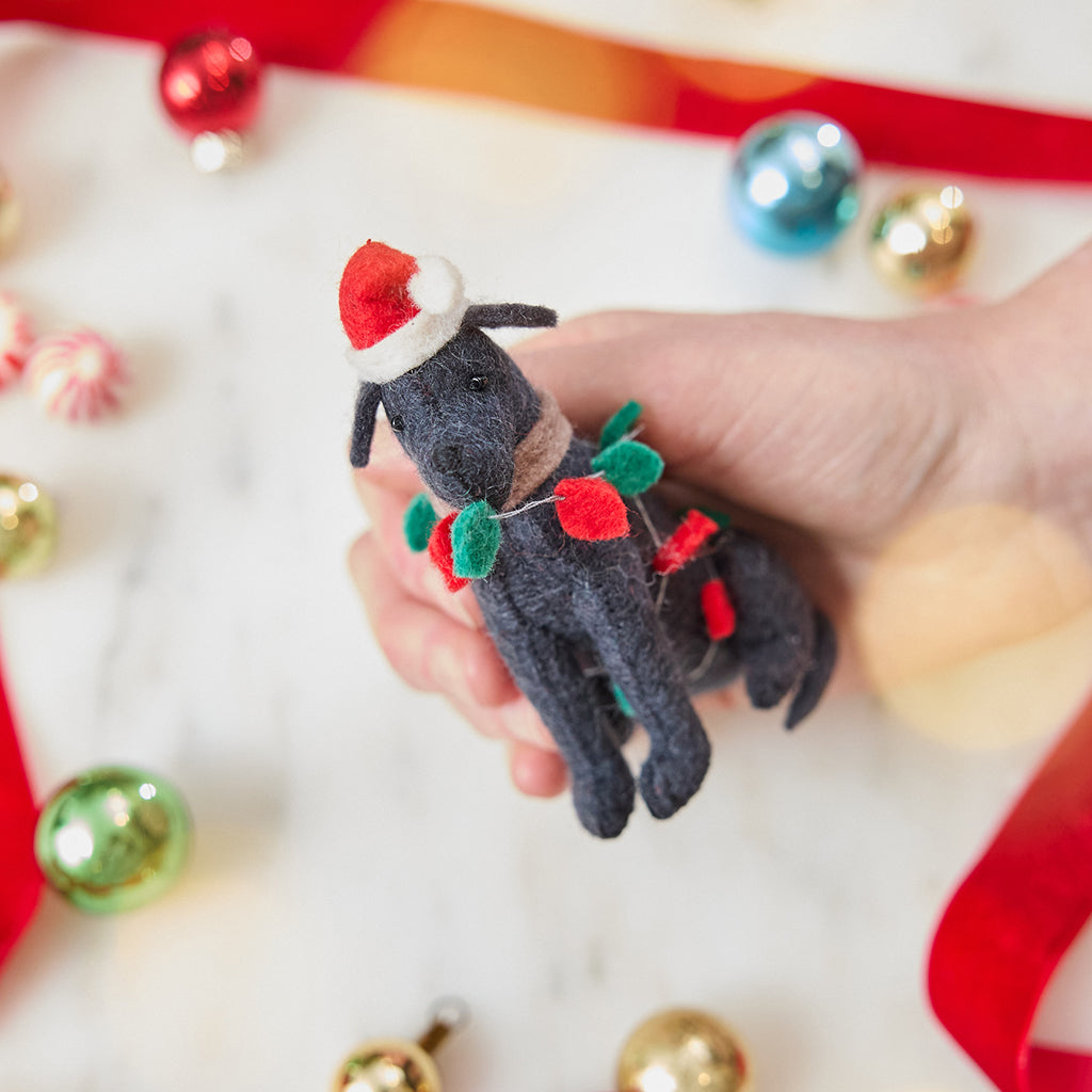 Craftspring’s Black Lab in Christmas Lights Ornament — felt dog wrapped in Christmas lights and wearing a Santa hat — displayed in front of hand over holiday baubles, capturing playful charm, festive warmth, and heirloom-quality Christmas gifting, ethically crafted by artisans using sustainable wool.