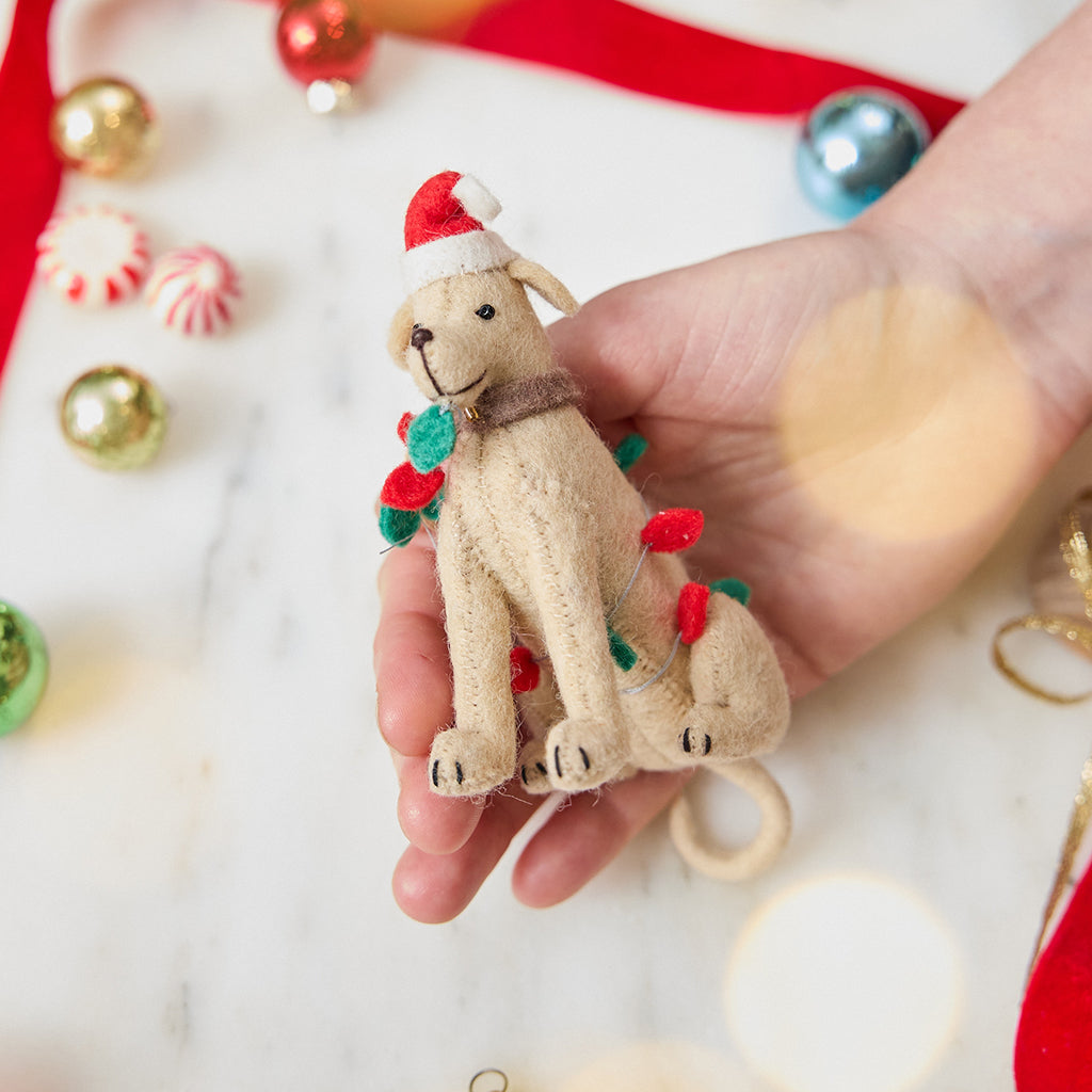 Craftspring’s Yellow Lab in Christmas Lights Ornament — felt dog wrapped in Christmas lights and wearing a Santa hat — held in hand over holiday baubles, capturing playful charm, festive warmth, and heirloom-quality Christmas gifting, ethically crafted by artisans using sustainable wool.