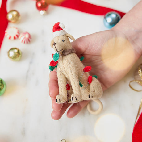 Craftspring’s Yellow Lab in Christmas Lights Ornament — felt dog wrapped in Christmas lights and wearing a Santa hat — held in hand over holiday baubles, capturing playful charm, festive warmth, and heirloom-quality Christmas gifting, ethically crafted by artisans using sustainable wool.