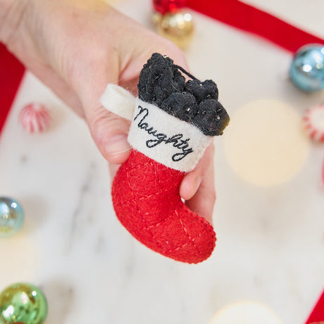 Craftspring’s Naughty Stocking with Coal Ornament — felt red stocking filled with felt coal and beads, embroidered “naughty” — held in hand over holiday baubles, capturing whimsical charm, festive warmth, and heirloom-quality Christmas gifting, ethically crafted by artisans using sustainable wool.