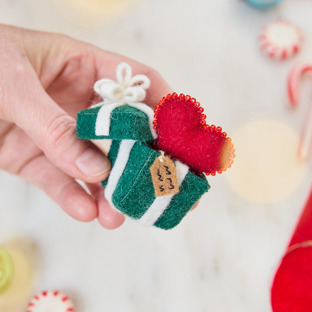 Craftspring’s Gift from the Heart Ornament — felt heart ornament with beaded and embroidered details including a gift tag — held in hand over candy canes and Christmas treats, adding festive charm and holiday gifting warmth, ethically crafted by artisans using sustainable wool.