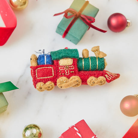 Craftspring’s Toy Train Ornament — felt red and green train with gold embroidery, carrying presents and a teddy bear — styled among holiday baubles and mini presents, evoking festive warmth, playful Christmas cheer, and heirloom-quality charm, ethically crafted by artisans using sustainable wool.