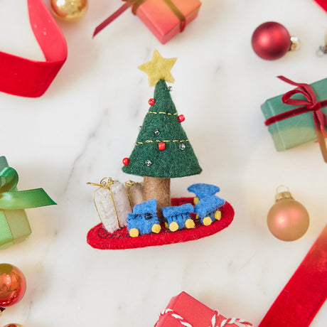 Craftspring’s Train Around the Christmas Tree Ornament — felt blue toy train wrapping around the base of a decorated Christmas tree with presents — styled among holiday baubles and mini presents, evoking playful Christmas cheer, festive warmth, and heirloom-quality gifting, ethically crafted by artisans using sustainable wool.
