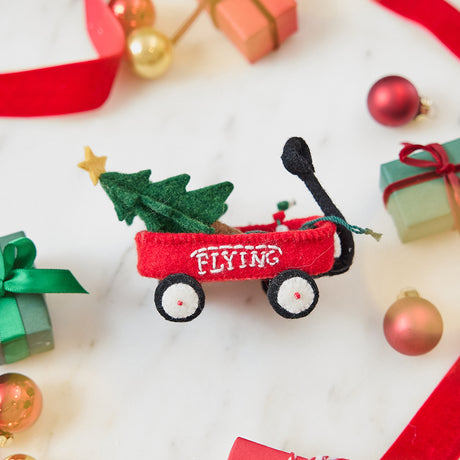 Craftspring’s Little Red Wagon with Tree Ornament — felt little red wagon carrying a Christmas tree and garland, with embroidered and beaded details — styled among holiday baubles and mini presents, evoking festive warmth, playful Christmas cheer, and heirloom-quality gifting, ethically crafted by artisans using sustainable wool.