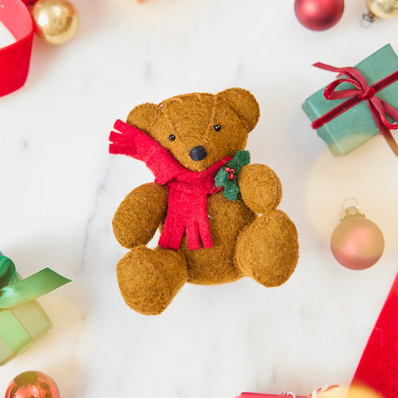 Craftspring’s Holiday Teddy Bear Ornament — felt vintage-style teddy bear wearing a red scarf with holly — styled among holiday baubles and mini presents, evoking festive warmth, playful Christmas cheer, and heirloom-quality gifting, ethically crafted by artisans using sustainable wool.