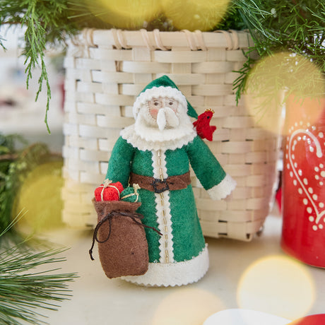 Craftspring’s Emerald Santa Mini Tree Topper — felt Santa in a green cloak with beaded details, holding a sack of presents and a bird on his shoulder — displayed in front of a basket full of evergreens, evoking festive holiday warmth, vintage charm, and heirloom-quality Christmas gifting, ethically crafted by artisans using sustainable wool.