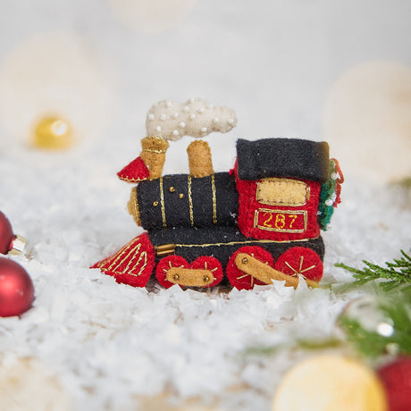 Craftspring’s Steam Engine Train Ornament — felt black and red train with gold embroidery and beading, fluffy steam, and a wreath on the back — displayed on snow with evergreens, capturing whimsical holiday spirit, cozy Christmas warmth, and heirloom-quality gifting, ethically crafted by artisans using sustainable wool.