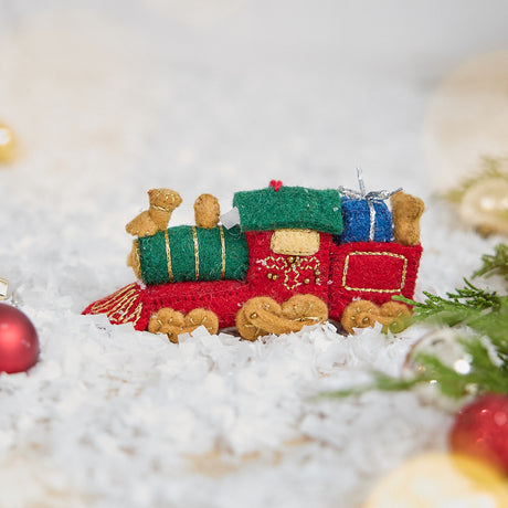 Craftspring’s Toy Train Ornament — felt red and green train with gold embroidery, carrying presents and a teddy bear — displayed on snow, evoking festive holiday charm, heirloom-quality warmth, and Christmas gifting joy, ethically crafted by artisans using sustainable wool.