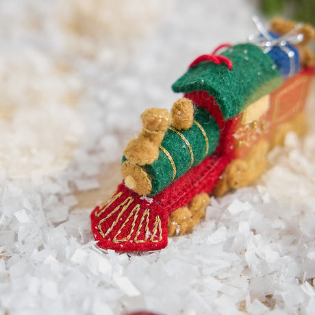 Craftspring’s Toy Train Ornament — felt red and green train with gold embroidery, intricately detailed with a team engine carrying presents and a teddy bear — displayed on snow showing the front of the train, capturing playful holiday spirit and heirloom-quality charm, ethically crafted by artisans using sustainable wool.