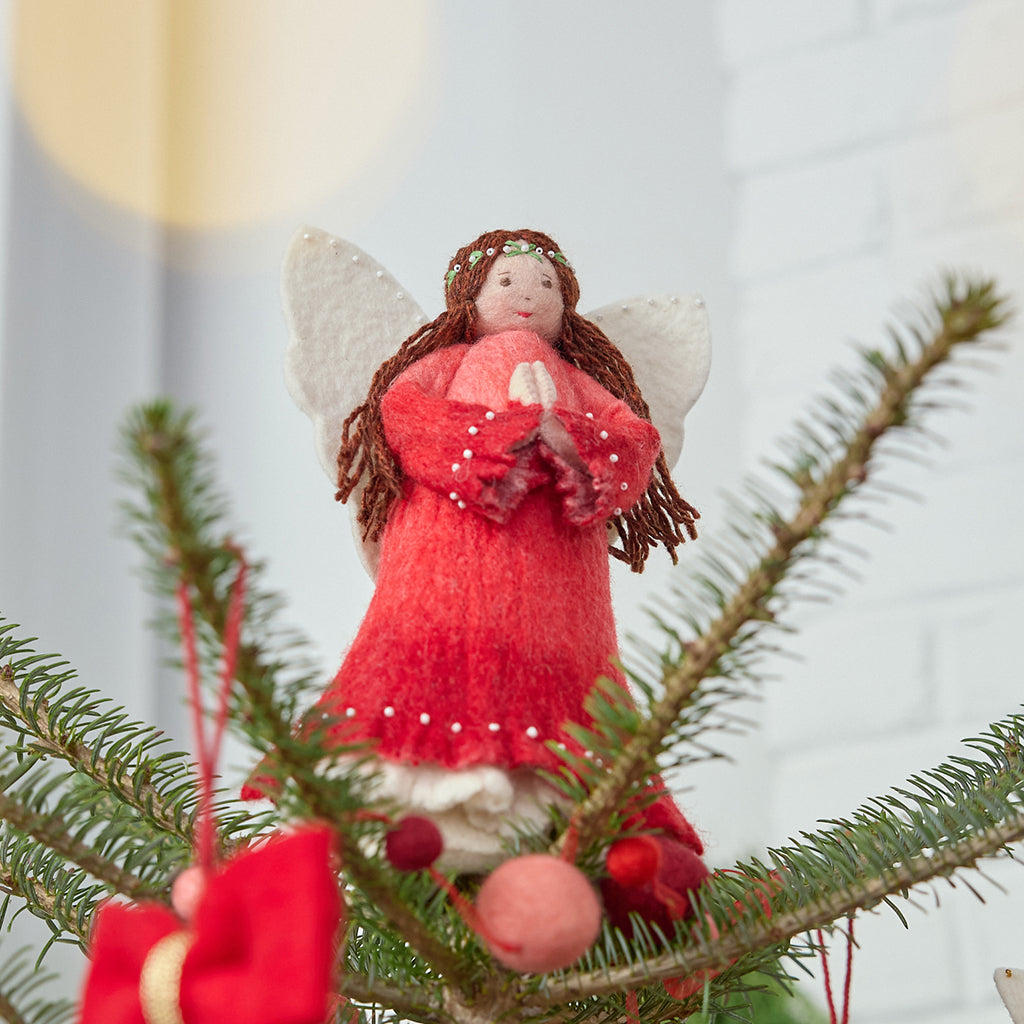 Craftspring’s Praying Angel Tree Topper - Light — felt angel wearing a red dress with white beads, embroidered and beaded wreath crown, and hands in prayer — displayed on top of a Christmas tree, evoking festive warmth, sweet holiday charm, and heirloom-quality Christmas gifting, ethically crafted by artisans using sustainable wool.