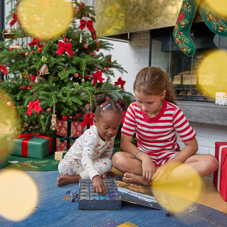 Craftspring’s Woodland Minis Advent Calendar — two girls enjoying the calendar by a tree with Craftspring ornaments, Woodland Tree Skirt, and stockings on the mantle, creating a cozy, gift-filled holiday scene, ethically crafted by artisans using sustainable wool.