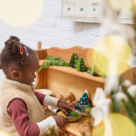 Craftspring’s Green Honeycomb Paper Mini Trees — Set of 5, displayed in a children’s play kitchen next to a Christmas tree, with a little girl playing with evergreens — showcasing the trees’ shades of green, gold sparkle, and unique shapes, handmade by artisans from recycled cotton waste for a cozy holiday scene.