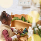 Craftspring’s Green Honeycomb Paper Mini Trees — Set of 5, displayed in a children’s play kitchen next to a Christmas tree, with a little girl playing with evergreens — showcasing the trees’ shades of green, gold sparkle, and unique shapes, handmade by artisans from recycled cotton waste for a cozy holiday scene.