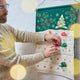 Craftspring’s Woodland Advent Calendar shown with a dad filling the pockets with mini presents — featuring intricate embroidered, appliqué, and beaded woodland details, a joyful holiday tradition ethically crafted by artisans using sustainable wool.