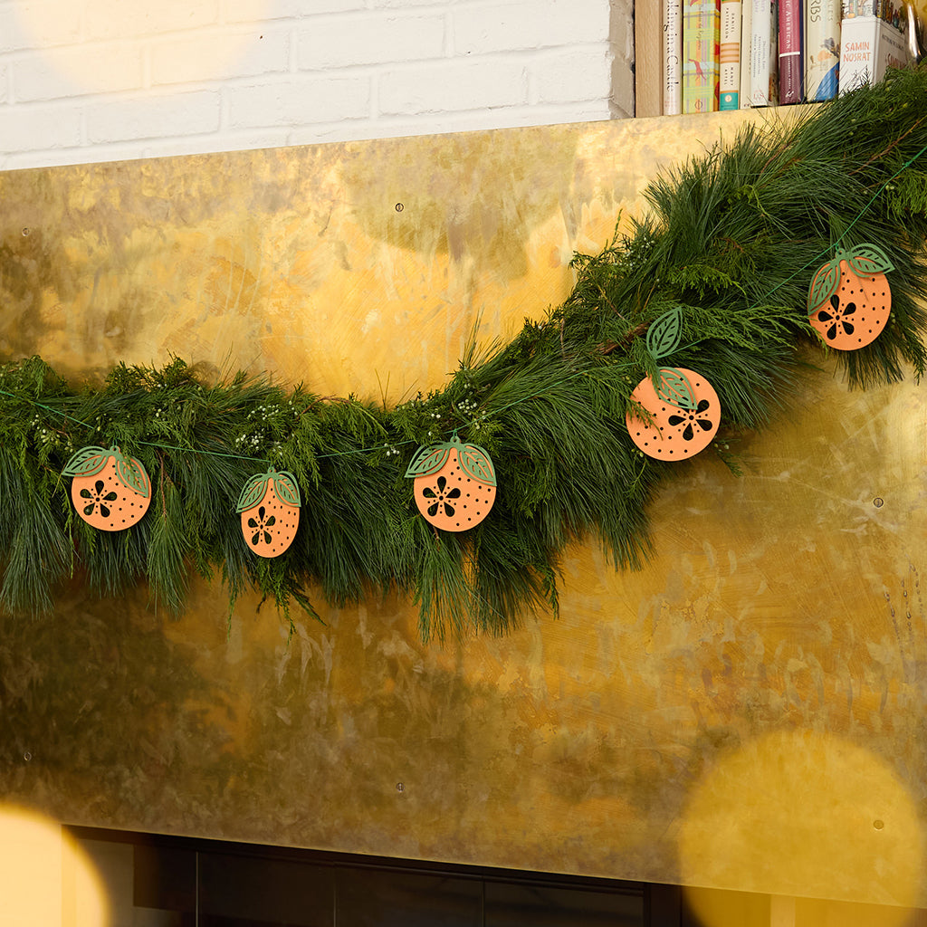 Craftspring’s Paper Clementine Garland — delicate handmade paper clementines with leaves and intricate cutout details, displayed on an evergreen garland draped across a mantle, ethically handmade by artisans from recycled cotton waste for festive Christmas, holiday, and Lunar New Year decor.