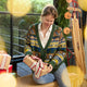 Craftspring’s NYC Holiday 100% Alpaca Sweater Cardigan — ultra-soft cardigan made of sustainably harvested baby alpaca wool, with intricate jacquard motifs of NYC icons like bagels, taxi cabs, and pigeons — heirloom-quality knitwear ethically crafted by artisans in Peru. A woman wears the sweater while holding a Christmas present in a festive holiday scene, embodying holiday warmth and gifting joy.