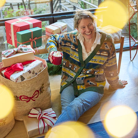 Craftspring’s NYC Holiday 100% Alpaca Sweater Cardigan — cozy heirloom cardigan crafted of 100% sustainably harvested baby alpaca wool with intricate jacquard NYC motifs, including taxis, pigeons, and bagels — ethically made by artisans in Peru. A woman wears the sweater while drinking hot cocoa in a festive holiday scene, capturing the warmth and spirit of Christmas gifting.
