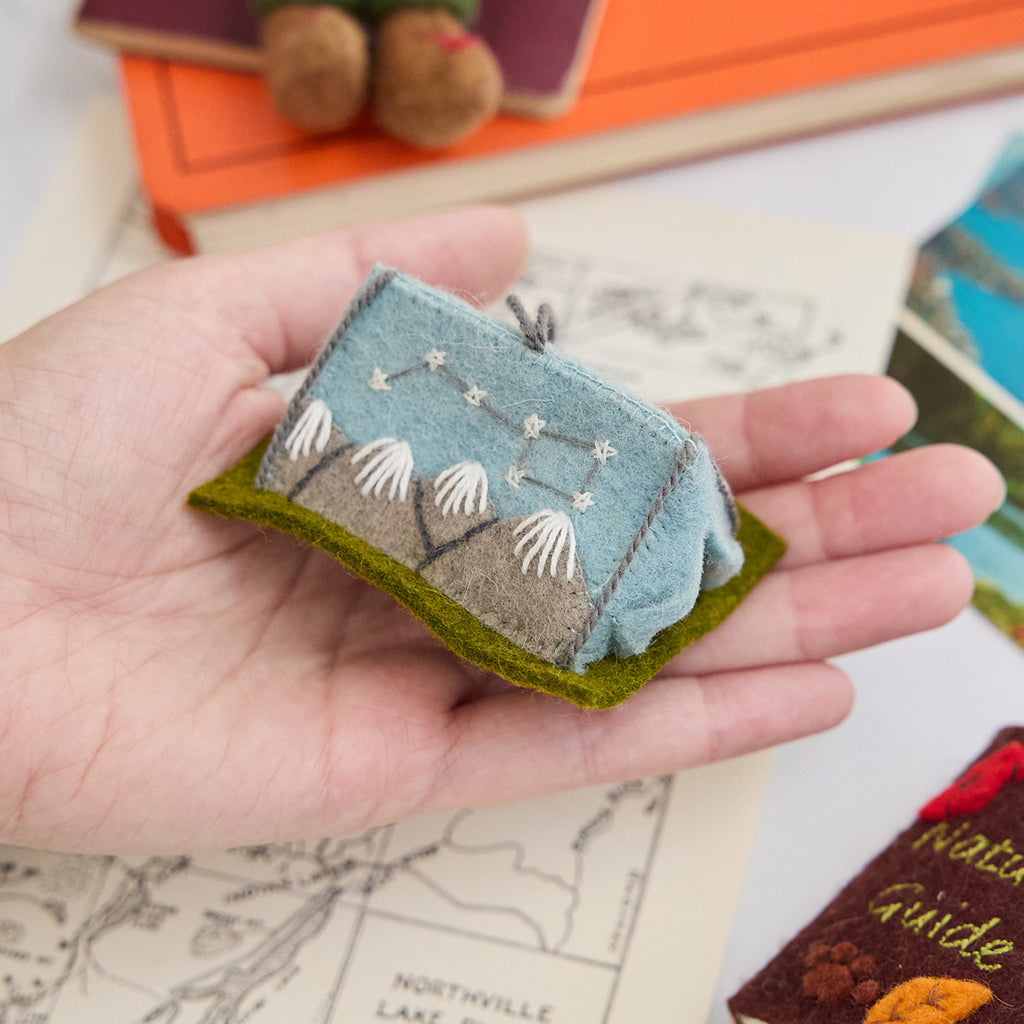 Craftspring’s handmade felt Home Under the Stars Tent ornament — applique and embroidered mountains with the Big Dipper on the side and a mini felt mattress inside the tent, held in a hand over a trail map to show its size, perfect for Christmas decorating or gifting, ethically crafted by artisans using sustainable wool.