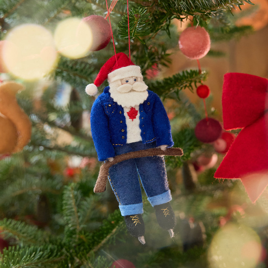 Craftspring’s Canadian Santa Ornament displayed on a Christmas tree — felt Santa wearing a denim jacket and jeans, ice skates, and holding a hockey stick, with a Canadian flag maple leaf on his shirt, adding festive warmth and whimsical holiday charm — heirloom-quality Christmas decor, ethically crafted by artisans using sustainable wool.
