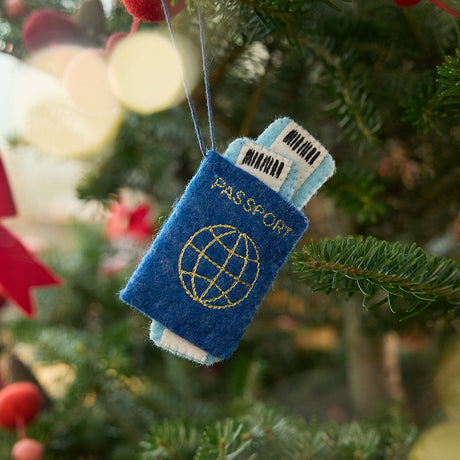 Craftspring’s Passport ornament — featuring applique and embroidered details with two plane tickets tucked inside and passport stamps on the back, displayed on a Christmas tree for a festive holiday scene, ethically crafted by artisans using sustainable wool.