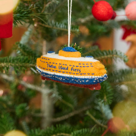 Craftspring’s Staten Island Ferry ornament — featuring intricate embroidered details and beaded waves on the iconic orange ferry, displayed on a Christmas tree for a festive holiday scene, ethically crafted by artisans using sustainable wool.
