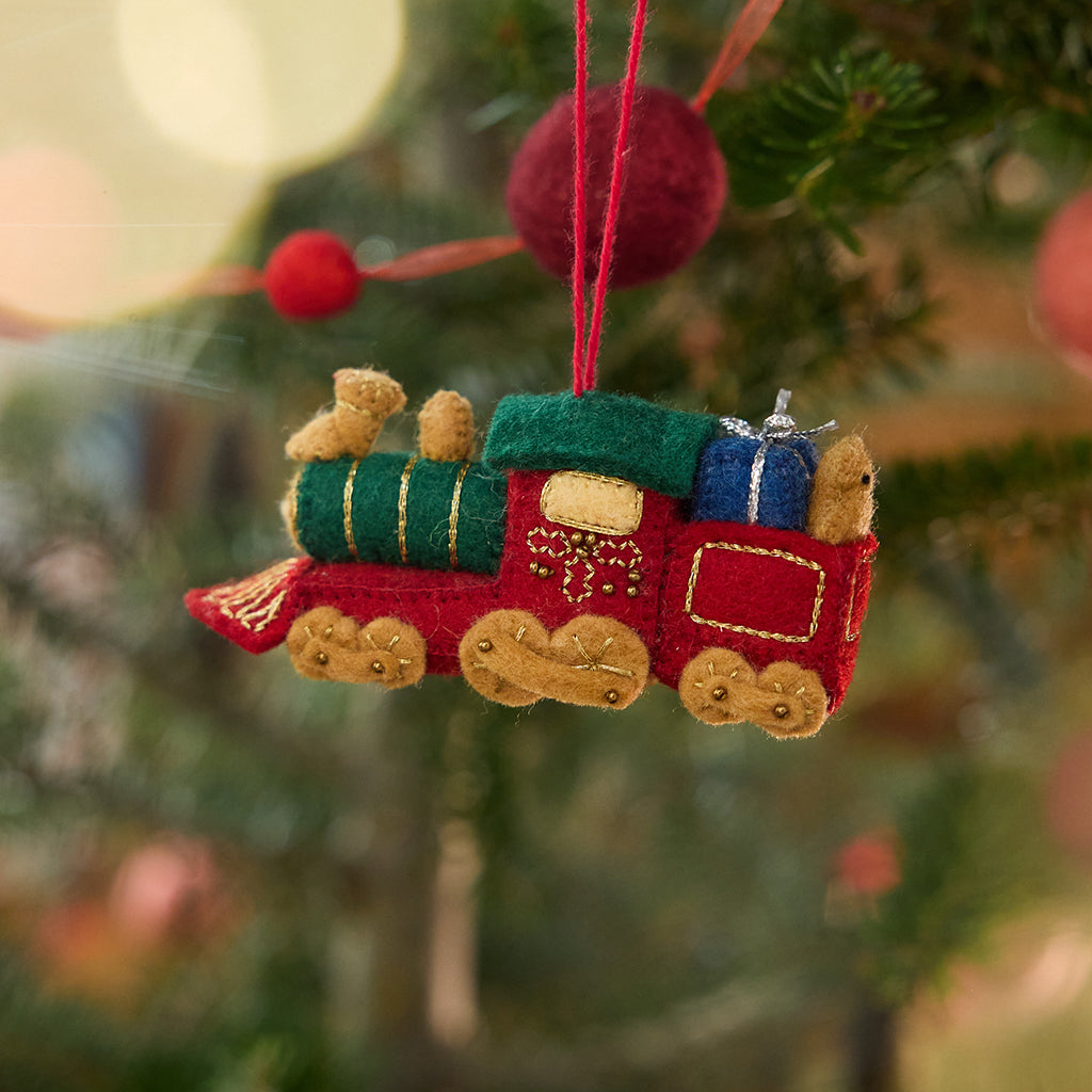 Craftspring’s Toy Train Ornament displayed on a Christmas tree — felt red and green train with gold embroidery, intricately detailed with a team engine carrying presents and a teddy bear, adding festive warmth and whimsical charm — heirloom-quality Christmas decor, ethically crafted by artisans using sustainable wool.