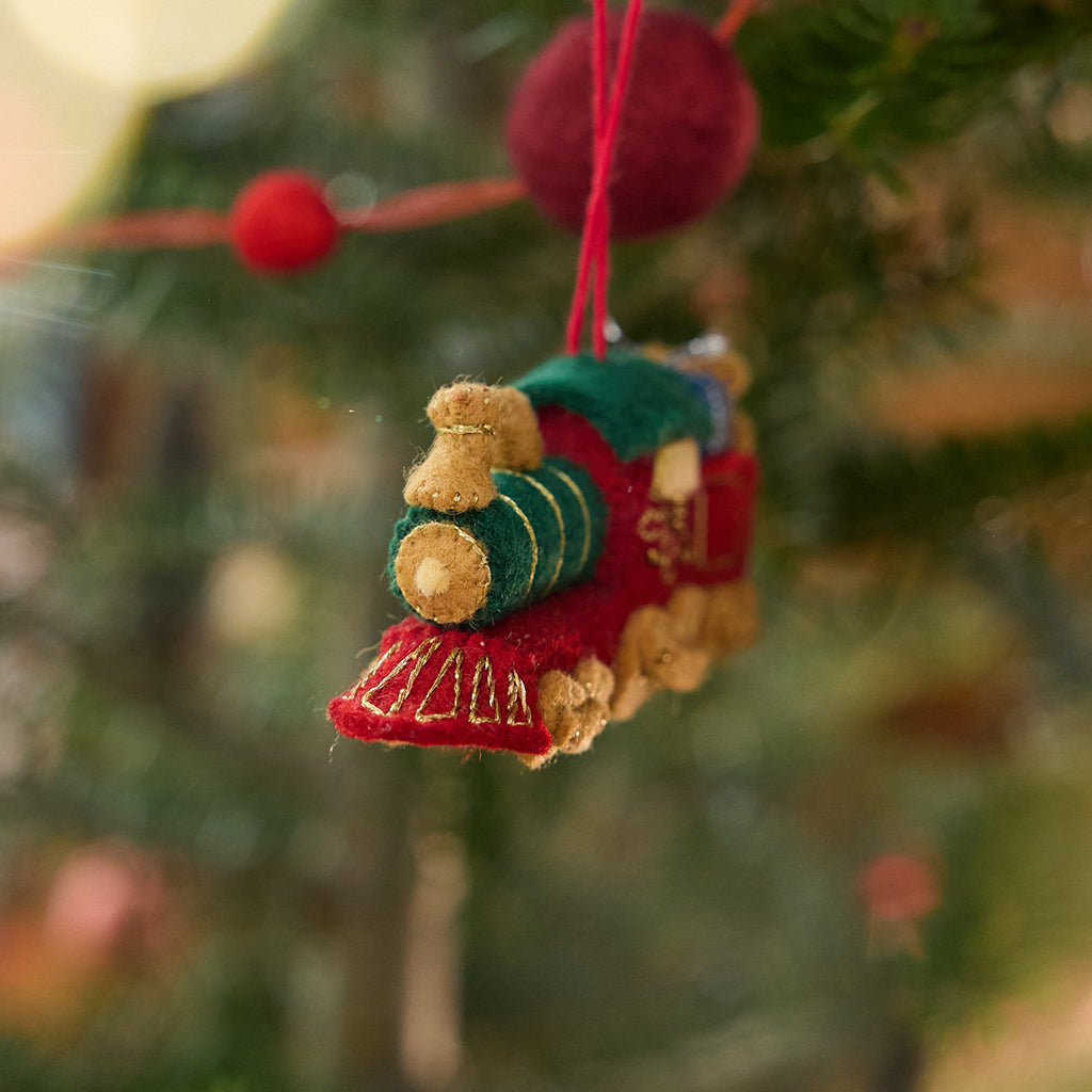 Craftspring’s Toy Train Ornament displayed on a Christmas tree — felt red and green train with gold embroidery, intricately detailed with a team engine carrying presents and a teddy bear, adding festive warmth and whimsical charm — heirloom-quality Christmas decor, ethically crafted by artisans using sustainable wool.