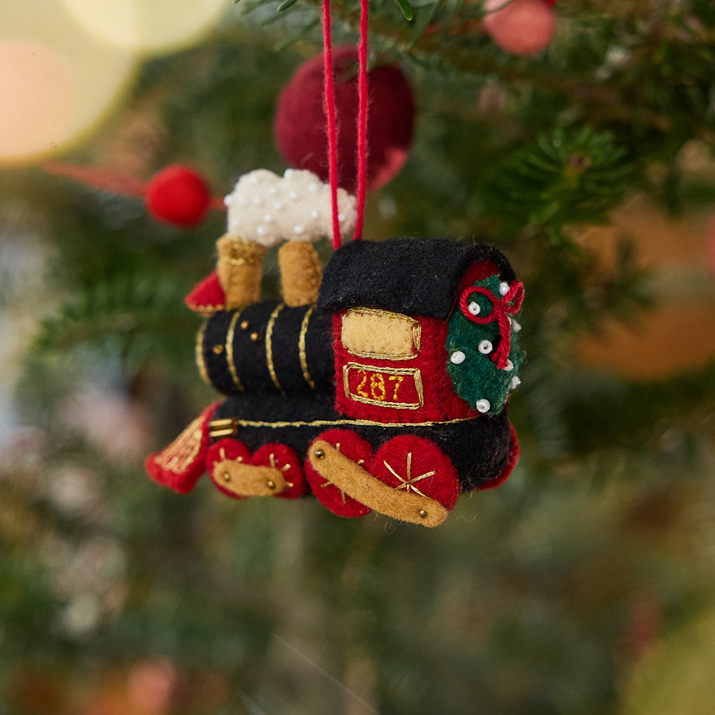 Craftspring’s Steam Engine Train Ornament displayed on a Christmas tree — felt black and red train with gold embroidery and beading, fluffy steam rising from the top, and a wreath on the back, adding festive warmth and whimsical charm — heirloom-quality Christmas decor, ethically crafted by artisans using sustainable wool.