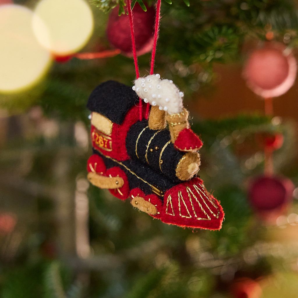 Craftspring’s Steam Engine Train Ornament displayed on a Christmas tree — felt black and red train with gold embroidery and beading, fluffy steam rising from the top, and a wreath on the back, adding festive warmth and whimsical charm — heirloom-quality Christmas decor, ethically crafted by artisans using sustainable wool.