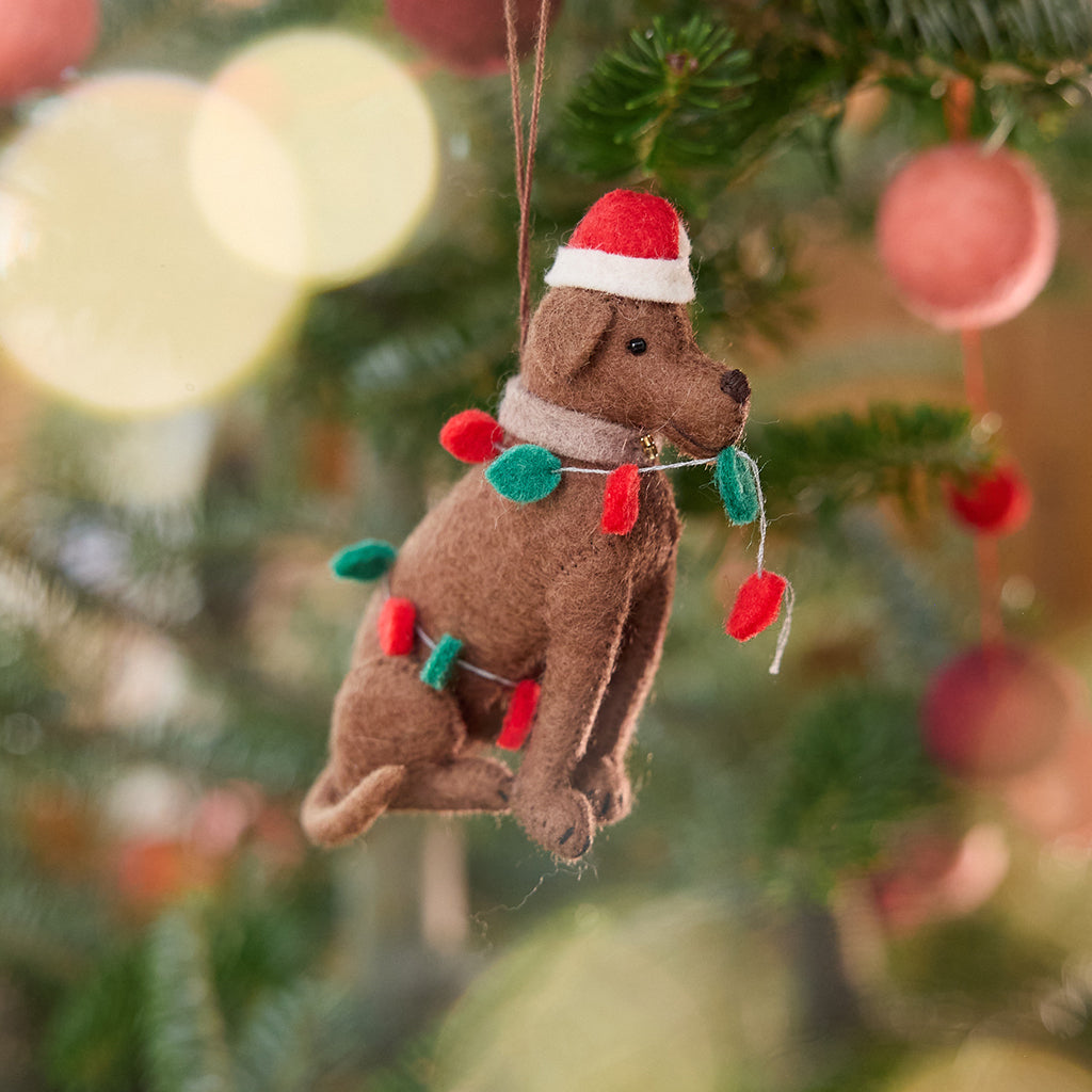 Craftspring’s Chocolate Lab in Christmas Lights Ornament displayed on a Christmas tree — felt dog wrapped in Christmas lights and wearing a Santa hat, adding playful holiday charm and festive warmth — heirloom-quality Christmas decor, ethically crafted by artisans using sustainable wool.