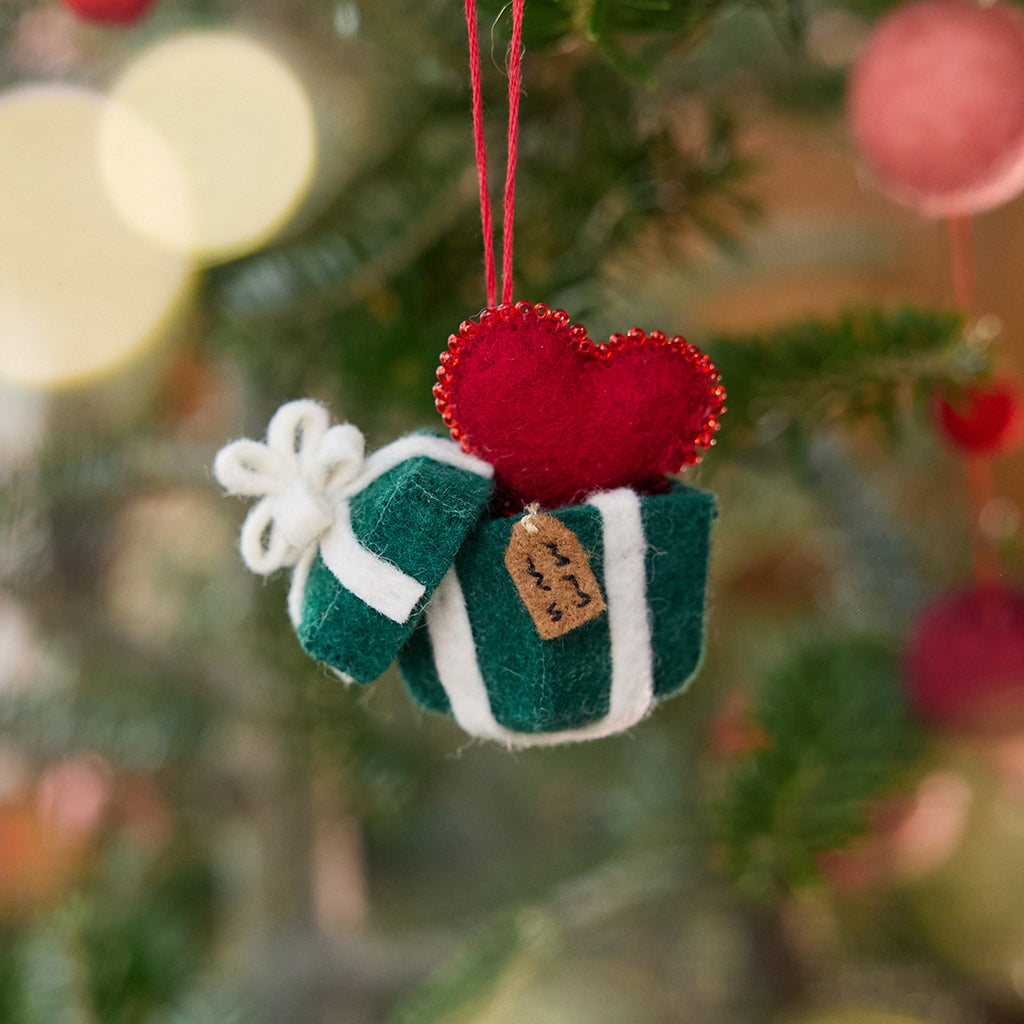 Craftspring’s Gift from the Heart Ornament displayed on a Christmas tree — felt ornament of a heart coming out of a present, with beaded and embroidered details, adding warm and festive holiday charm, ethically crafted by artisans using sustainable wool.