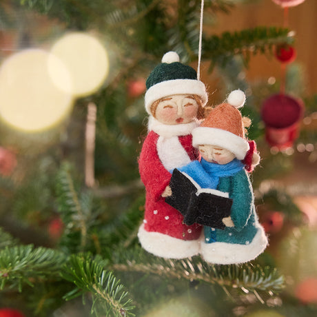 Mom and Child Carolers Ornament from Craftspring — handmade felt holiday ornament showing a mother and child singing together and sharing a Noel book, hanging on a Christmas tree with intricate applique and embroidered snow details, ethically crafted by artisans using sustainable wool.