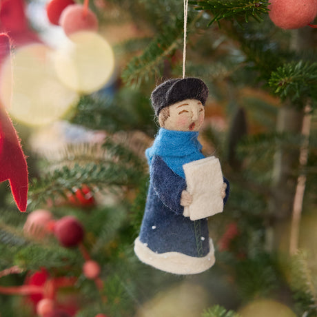 Caroler Holding Sheet Music Ornament from Craftspring — handmade felt holiday ornament with intricate applique and embroidered snow, holding sheet music and hanging on a Christmas tree, ethically crafted by artisans using sustainable wool.