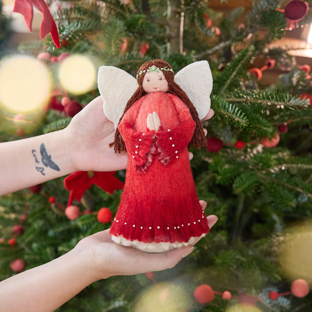 Craftspring’s Praying Angel Tree Topper - Light — felt angel in a red dress with white beads, with embroidered and beaded wreath crown and hands in prayer — held in hand in front of a Christmas tree, capturing cozy holiday magic, whimsical charm, and heirloom-quality gifting, ethically crafted by artisans using sustainable wool.