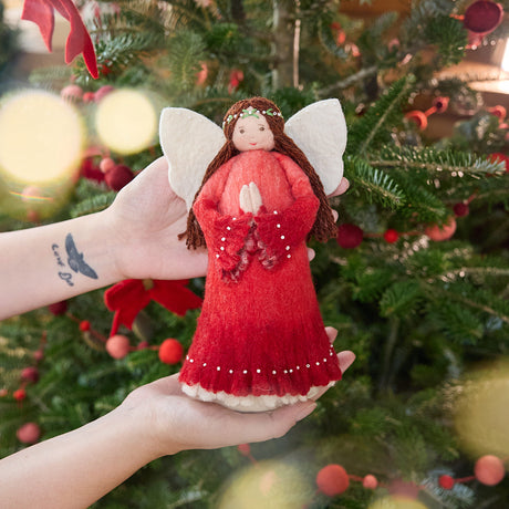 Craftspring’s Praying Angel Tree Topper - Light — felt angel in a red dress with white beads, with embroidered and beaded wreath crown and hands in prayer — held in hand in front of a Christmas tree, capturing cozy holiday magic, whimsical charm, and heirloom-quality gifting, ethically crafted by artisans using sustainable wool.