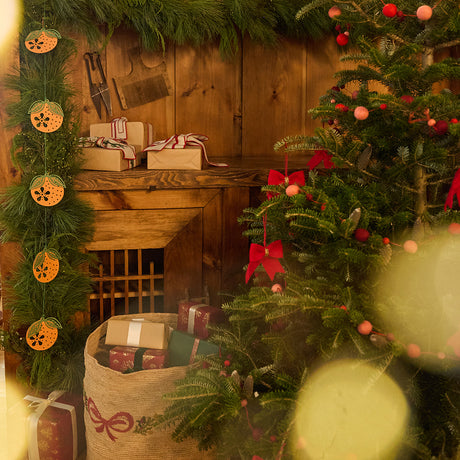 Craftspring holiday scene with a Christmas tree decorated with red bow and red ball garlands, surrounded by presents, and an evergreen garland adorned with Craftspring’s Paper Clementine Garland — festive Christmas, holiday, and Lunar New Year decor, ethically handmade by artisans from recycled cotton waste.