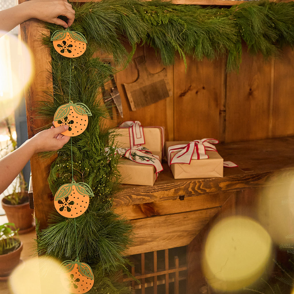 Craftspring’s Paper Clementine Garland — hands adjusting the handmade paper garland as it’s displayed on an evergreen garland, a festive and sustainable Christmas and Lunar New Year accent, ethically handmade by artisans from recycled cotton waste.