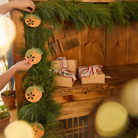 Craftspring’s Paper Clementine Garland — hands adjusting the handmade paper garland as it’s displayed on an evergreen garland, a festive and sustainable Christmas and Lunar New Year accent, ethically handmade by artisans from recycled cotton waste.
