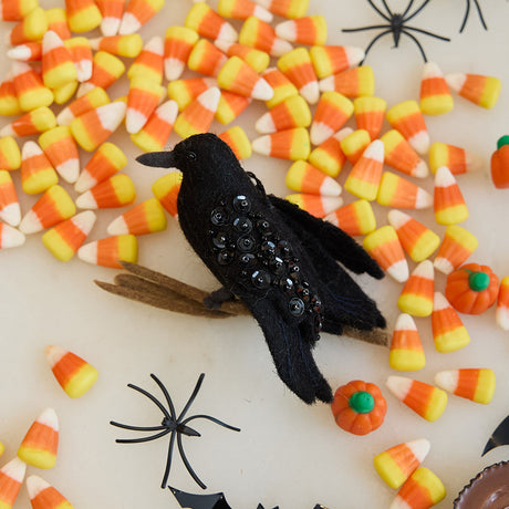 Craftspring’s handmade felt Crow ornament — featuring shimmering beaded wings and perched on a branch, displayed with Halloween candy for a festive autumn scene, perfect for Halloween, harvest, and holiday gifting, ethically crafted by artisans using sustainable wool.