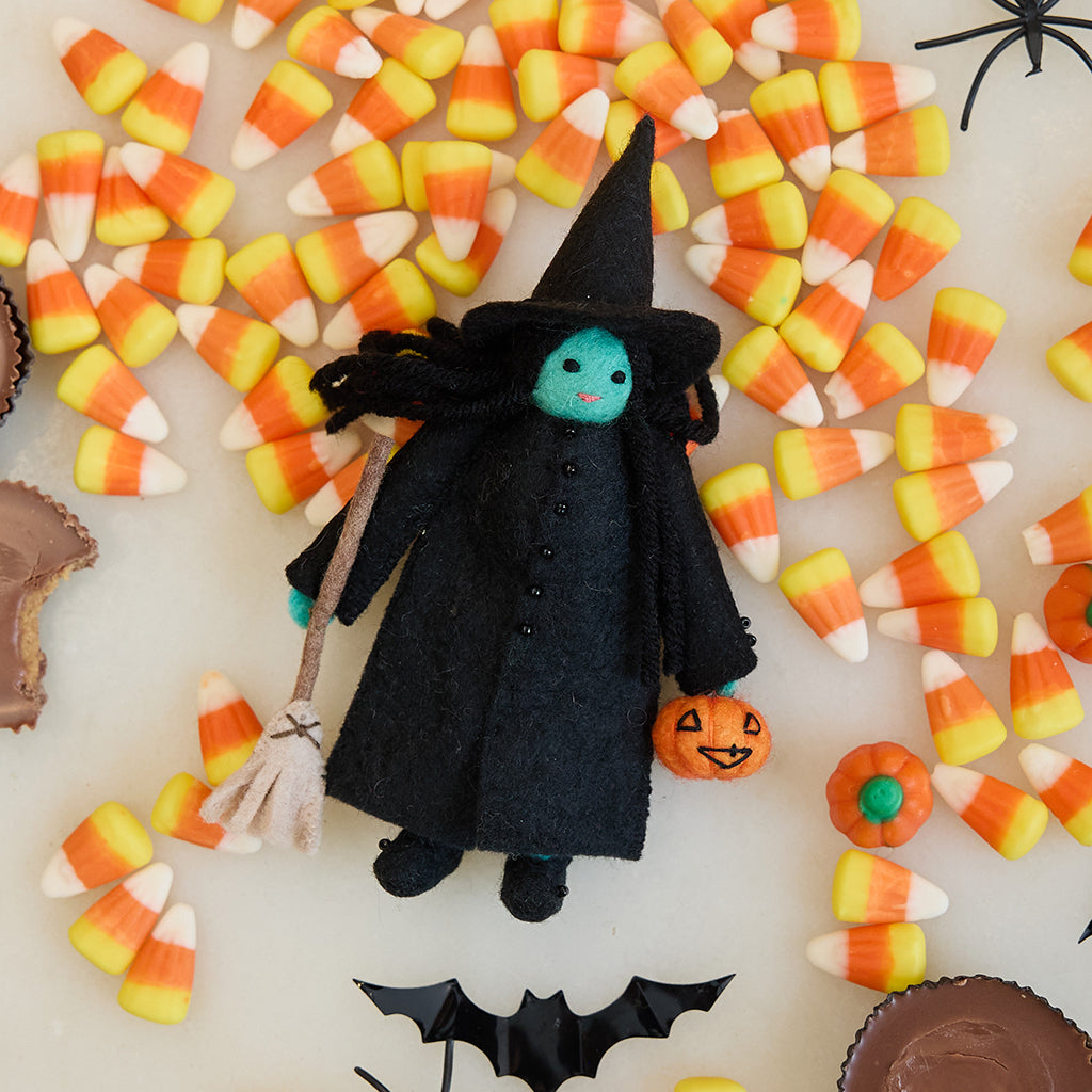 Craftspring’s Wicked Witch Trick-or-Treater ornament — with green skin, a witch’s hat, broom, and pumpkin basket — displayed with candy corn for a playful Halloween scene, perfect for spooky decorating and holiday gifting, ethically crafted by artisans using sustainable wool.