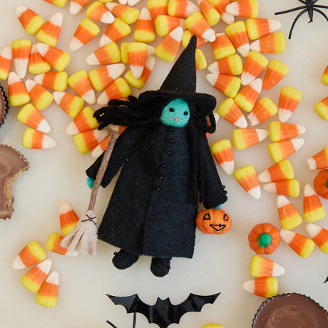 Craftspring’s Wicked Witch Trick-or-Treater ornament — with green skin, a witch’s hat, broom, and pumpkin basket — displayed with candy corn for a playful Halloween scene, perfect for spooky decorating and holiday gifting, ethically crafted by artisans using sustainable wool.