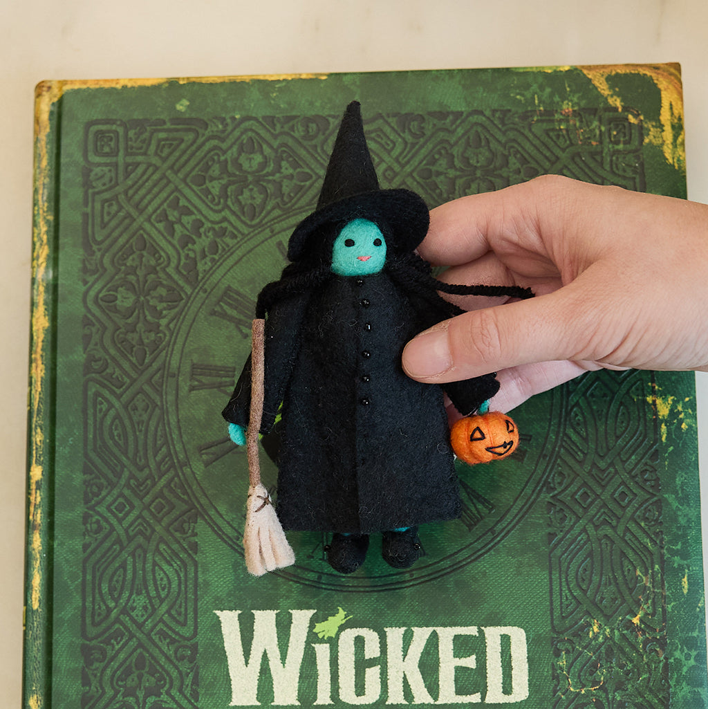 Craftspring’s Wicked Witch Trick-or-Treater ornament — with green skin, a witch’s hat, broom, and pumpkin basket — displayed on the book Wicked for a festive Halloween scene, perfect for spooky decorating and holiday gifting, ethically crafted by artisans using sustainable wool.